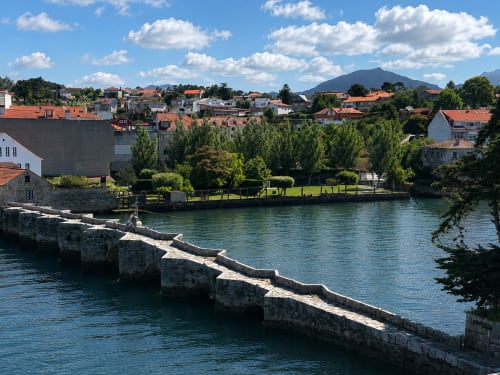 Puente histórico de piedra cruzando río turquesa en Baiona, España con casas de techos coloridos y montañas