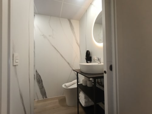 Baño moderno en El Rincón de Sevilla con baldosas de mármol blanco, lavamanos redondo, tocador negro, inodoro y espejo iluminado