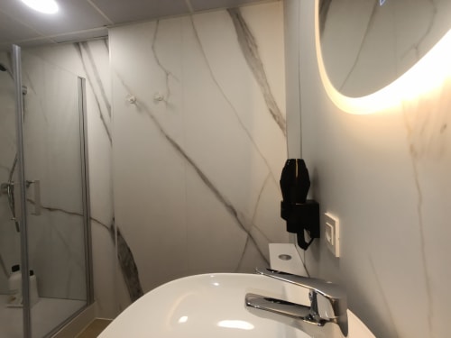 Baño moderno con paredes de mármol, espejo redondo con iluminación ambiental, lavamanos blanco y ducha en El