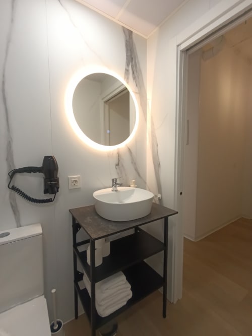 Baño moderno con espejo circular iluminado, lavamanos redondo blanco sobre estructura negra, pared de mármol en El Rincón de Sevilla