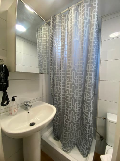 Baño moderno con lavabo de pedestal blanco, espejo, cortina de ducha con patrón geométrico y paredes con azulejos blancos en El Rincón de