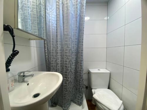 Baño limpio con azulejos blancos, lavabo con espejo, inodoro y cortina de ducha gris estampada en El Rincón de Sevilla