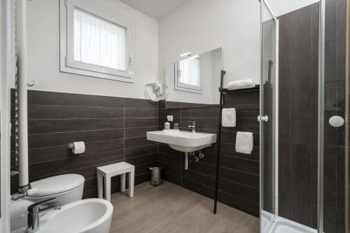 Bagno moderno e accessibile dell'Hotel Villa Mary con pareti piastrellate scure, lavabo bianco, toilette, bidet e doccia a filo pavimento