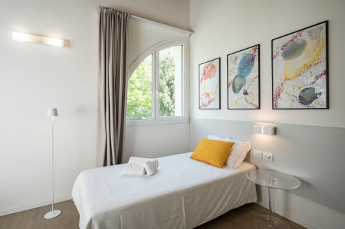 Camera da letto moderna a Hotel Villa Mary con vista sul giardino, letto bianco, cuscino giallo, opera costiera incorniciata e luce naturale