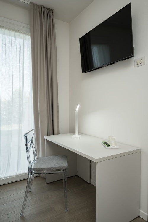 Moderna camera d'albergo con zona lavoro, scrivania bianca, sedia grigia, TV a muro e vista sul giardino attraverso le tende