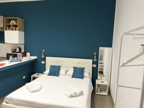 Camera moderna presso Harmony House, Bari con pareti azzurro teal, letto crema, cuscini blu e angolo cottura.