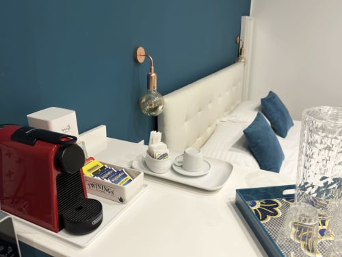 Dettaglio camera moderna presso l'Harmony House Bari con comodino bianco, macchina da caffè rossa, tè Twinings, lampada in rame da parete, e