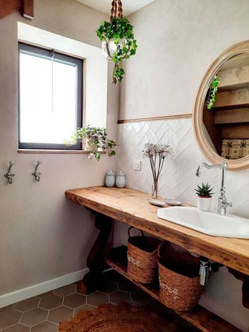 Rustic bathroom vanity with wooden countertop, white sink, round mirror, hanging plants, and woven baskets at Le Petit Bégou
