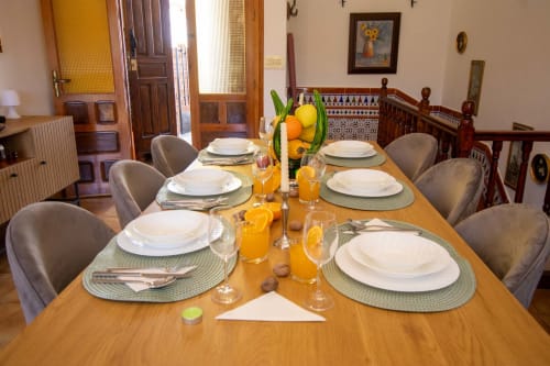 Mesa de comedor elegantemente preparada con cubiertas, jugo de naranja y flores amarillas en la cabaña La Casa de Tita Clota en Setenil de