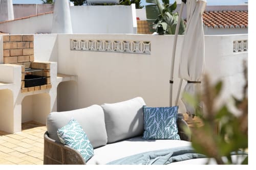 Cozy villa patio with wicker lounge chair, blue patterned pillows, white cushions, brick fireplace, and potted plants at