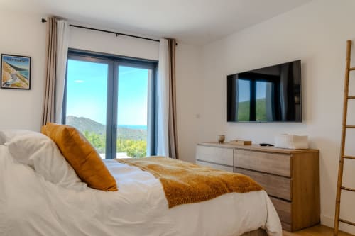 Modern bedroom at Villa Roya in Saint-Florent with sea view, white bed, orange pillows, wall-mounted TV, and terrace doors
