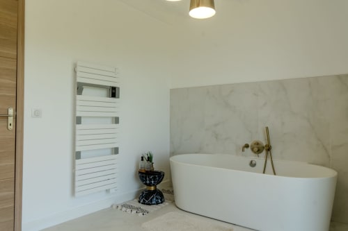 Modern bathroom at Villa Roya in Saint-Florent with oval white bathtub, marble accent wall, heated towel rail, and brass