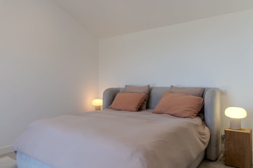 Modern bedroom at Villa Roya with gray upholstered bed, terracotta pillows, and ambient bedside lighting in Saint-Florent