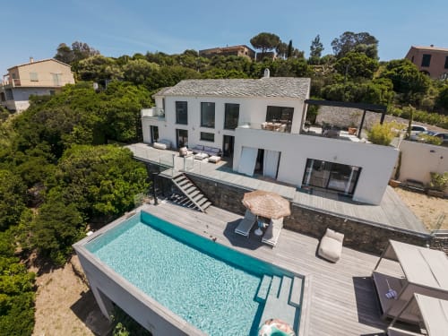 Villa Roya in Saint-Florent: modern white villa with turquoise pool, wooden deck, and surrounding greenery under blue sky
