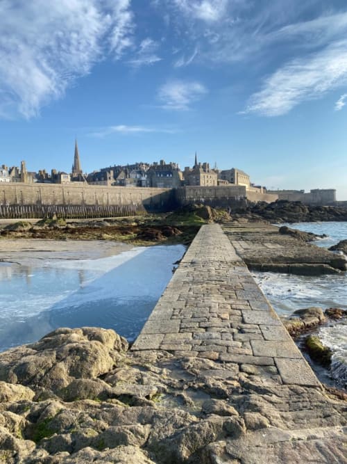 Chaussée en pierre menant à la cité médiévale fortifiée de Saint-Benoît-des-Ondes avec ses clochers, côte rocheuse et ciel bleu