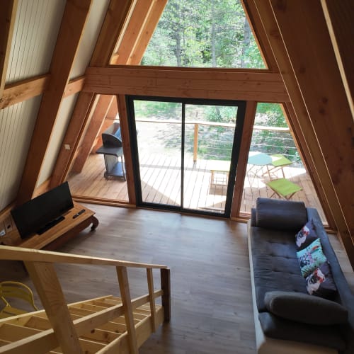 Intérieur de chalet en A moderne avec poutres en bois, canapé noir, bureau et portes vitrées donnant sur terrasse avec vue forêt à La Font