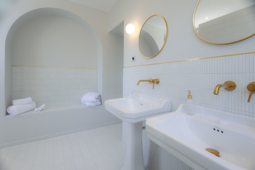 Salle de bains moderne à L'Ode à Pierre à Dax avec vasques doubles, robinetterie dorée, carrelage blanc et baignoire dans niche voûtée