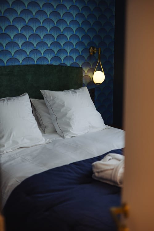 Chambre élégante à L'Ode à Pierre à Dax : oreillers blancs, tête de lit foncée, papier peint à motifs bleus et suspension en laiton