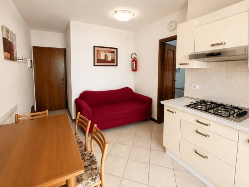Cucina e soggiorno dell'Apartment Timavo all'Hotel Anthea di Lignano Sabbiadoro con cucina a gas, divano rosso e tavolo da pranzo