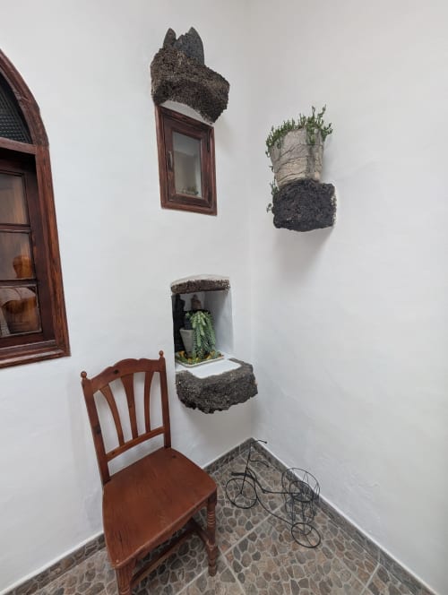 Rincón de villa en Lanzarote con silla de madera, macetas de roca volcánica con plantas verdes, marco de ventana de madera y suelo de piedra