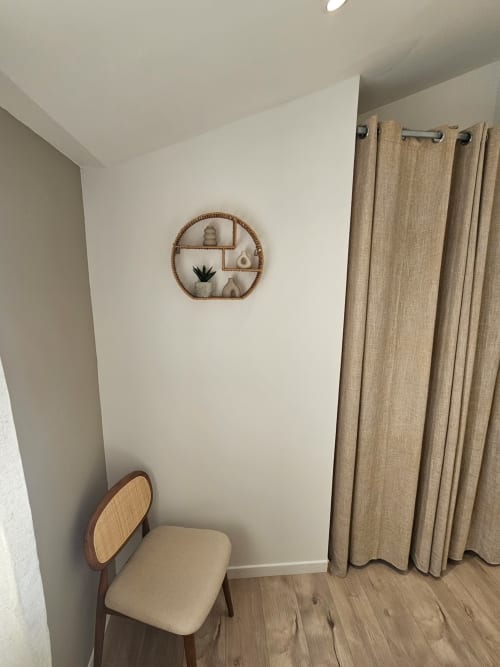 Coin minimaliste de la villa Altezza à Brando avec étagère murale circulaire en osier, fauteuil beige et rideaux neutres