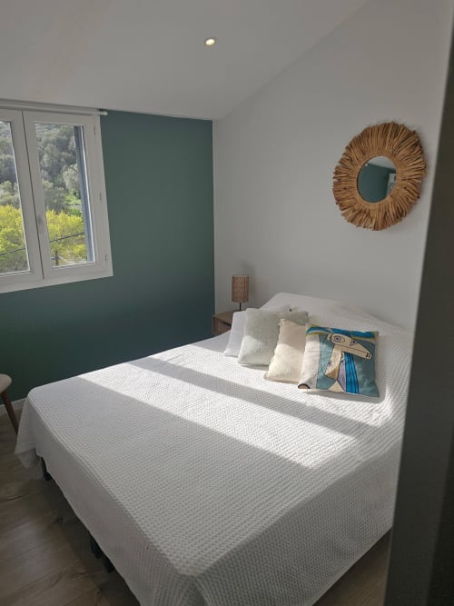 Chambre lumineuse avec lit blanc, oreillers crème, miroir tissé décoratif et mur d'accent vert à la villa Altezza à Brando