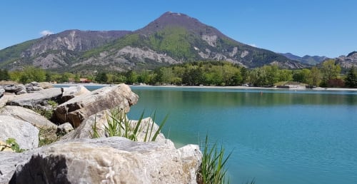 Lac turquoise avec rochers au premier plan, chaîne de montagnes en arrière-plan et arbres près du Petit Bégou à Saint-Auban-d'Oze