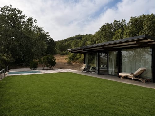 Casa de piscina moderna de negro con grandes ventanales, tumbonas de madera, jardín cuidado y colinas boscosas en El Escondite De Pedro
