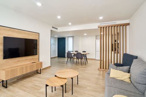 Moderna sala de estar diáfana en apartamento Tr3sor 11, Calella, con mueble de TV en madera, sofá gris, mesa de comedor y acabados de madera