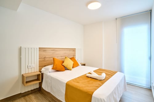 Moderno dormitorio en apartamento Tr3sor 11, Calella, con cama de madera, almohadas naranjas y amarillas, ropa de cama blanca y vidrios corredizos