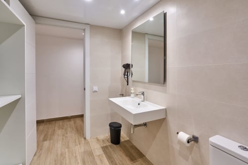 Baño moderno en apartamento Tr3sor Homes, Calella, con lavabo blanco, espejo, inodoro y piso de madera