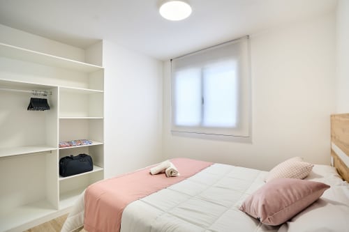 Habitación luminosa en Tr3sor Homes Calella con estanterías blancas, ropa de cama rosa, cama de madera y luz natural de la ventana