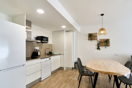 Moderna cocina y comedor del apartamento Tr3sor 13 en Calella con muebles blancos, mesa de madera e iluminación colgante