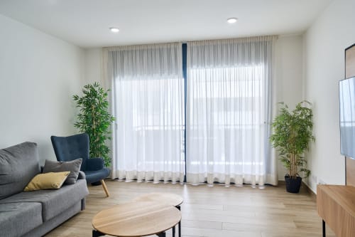 Moderno salón en Tr3sor 21 de Calella con sofá gris, silla azul, suelo de madera y cortinas blancas