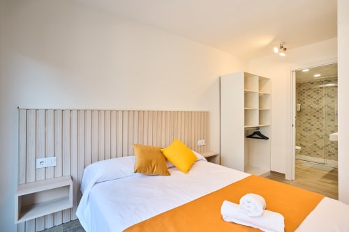 Moderno dormitorio en Tr3sor Homes Calella con cama queen, almohadas amarillas, baño en suite y almacenamiento integrado.