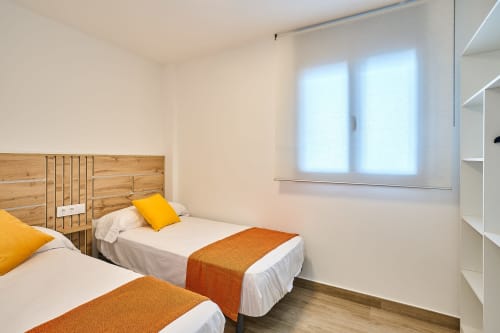 Dormitorio doble en apartamento Tr3sor Homes, Calella, con cabeceros de madera, acentos naranjas y luminosas vistas desde la ventana