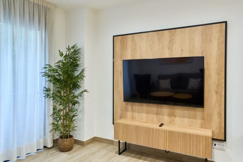Moderno salón en Tr3sor Homes Calella con TV de pared, mueble de madera y planta de bambú junto a cortinas translúcidas