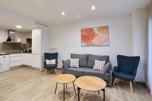 Sala moderna en Tr3sor Homes Calella con sofá gris, sillones azul marino, mesas de madera y cocina abierta