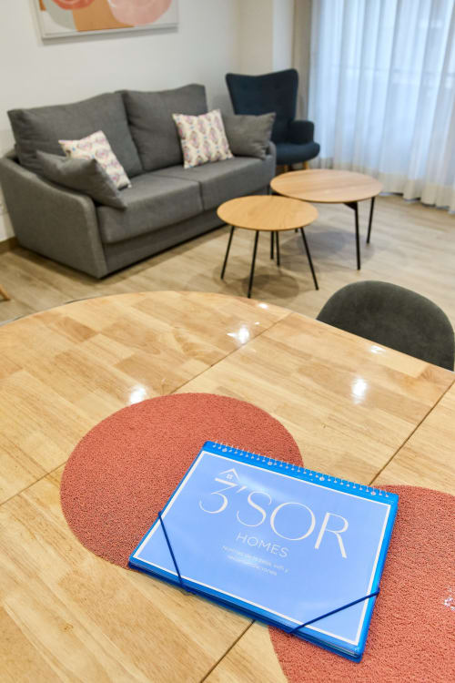 Folleto Tr3sor Homes azul sobre alfombra coral en salón moderno con sofá gris y mesas de madera, Calella