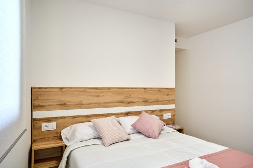 Moderno dormitorio en Tr3sor Homes Calella con cabecero de madera, ropa de cama blanca, almohadas rosas y mesitas incorporadas
