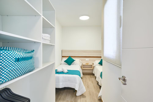 Dormitorio moderno en Tr3sor Homes, Calella con dos camas, detalles turquesas, suelo de madera y estanterías integradas.