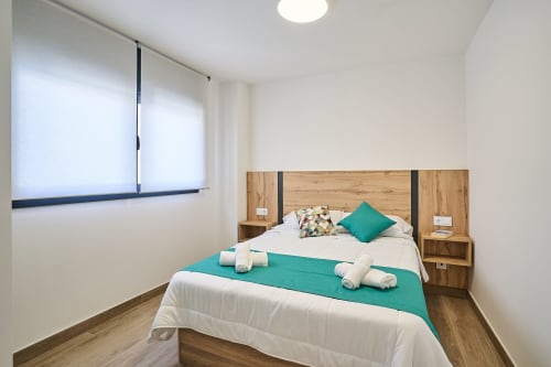 Dormitorio moderno en Tr3sor Homes Calella con cabecero de madera, detalles turquesas, ropa de cama blanca, toallas enrolladas y luz natural