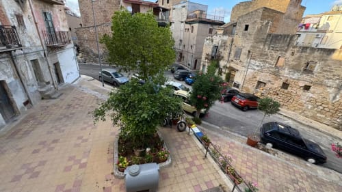 Vista dal balcone della storica piazza Santa Flavia con auto parcheggiate, edifici in pietra e vegetazione verde da Solunto Home