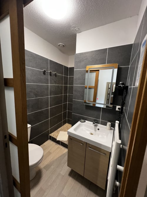 Bagno moderno dell'Hôtel les Alizés con doccia con piastrelle nere, lavabo bianco, wc e mobile in legno a Puget-Théniers