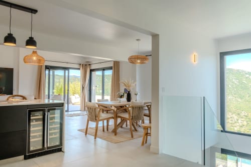 Salle à manger contemporaine à la Villa Roya à Saint-Florent avec chaises en bois, luminaires suspendus et vues panoramiques sur les montagnes