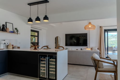 Villa Roya Saint-Florent, salon ouvert avec cuisine noire, luminaires suspendus, mur TV et terrasse vue mer