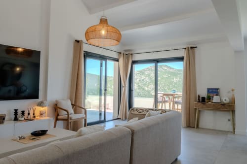 Salon élégant de la Villa Roya à Saint-Florent avec vue sur les montagnes, canapé beige, luminaire suspendu tissé et portes-fenêtres