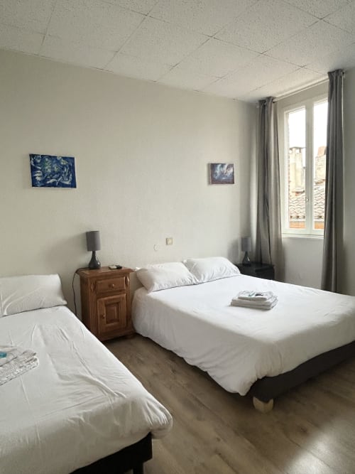Chambre quad lumineuse à l'hôtel BDM à Montastruc-la-Conseillère avec deux lits, table de nuit en bois et vue sur la ville