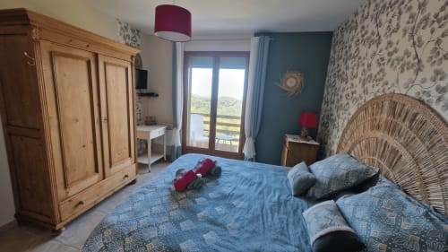 Chambre lumineuse à Mare e Monti Vico avec literie bleue, armoire en bois, tête de lit en rotin et vue sur les montagnes depuis le balcon