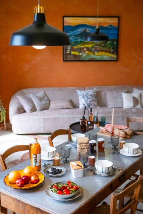 Table de petit-déjeuner dressée au Jardin Secret, Grignan, avec fruits frais, pâtisseries et salon aux murs orangés